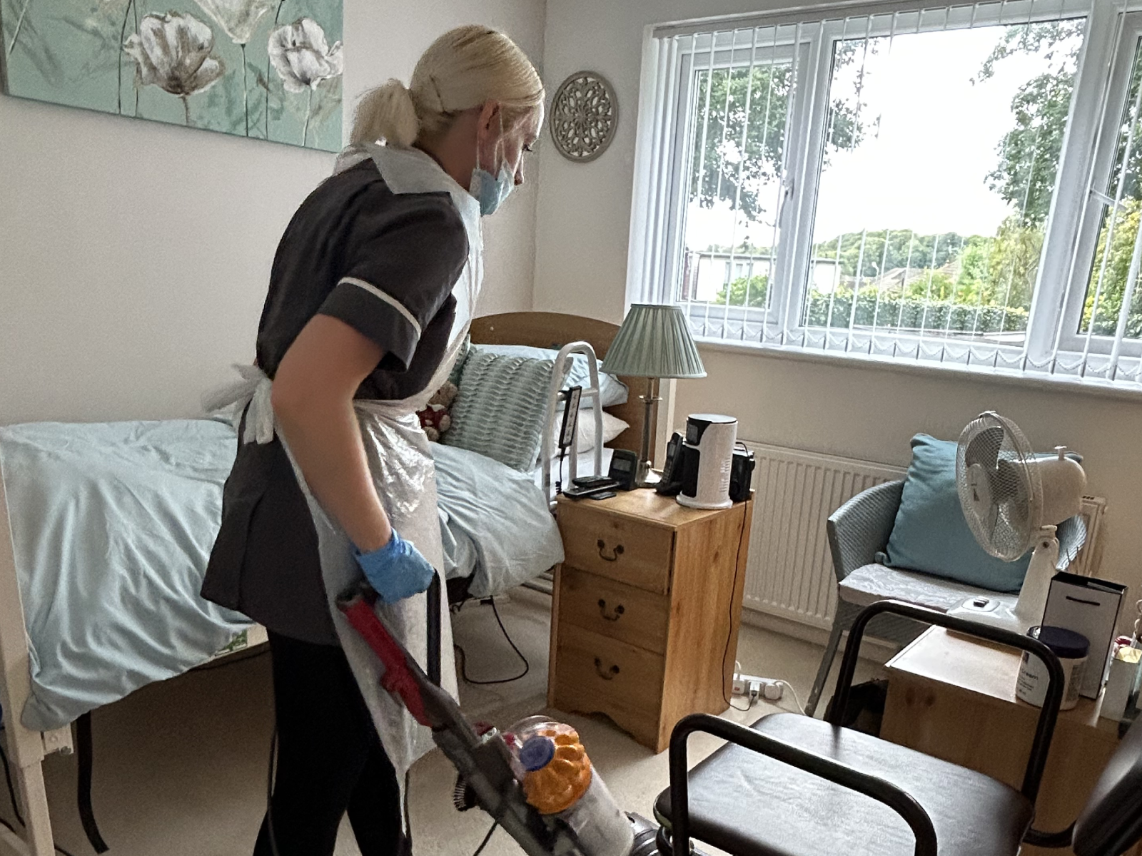 A caregiver cleaning a room while wearing gloves and a mask, highlighting the importance of jobs in care for maintaining hygiene and comfort