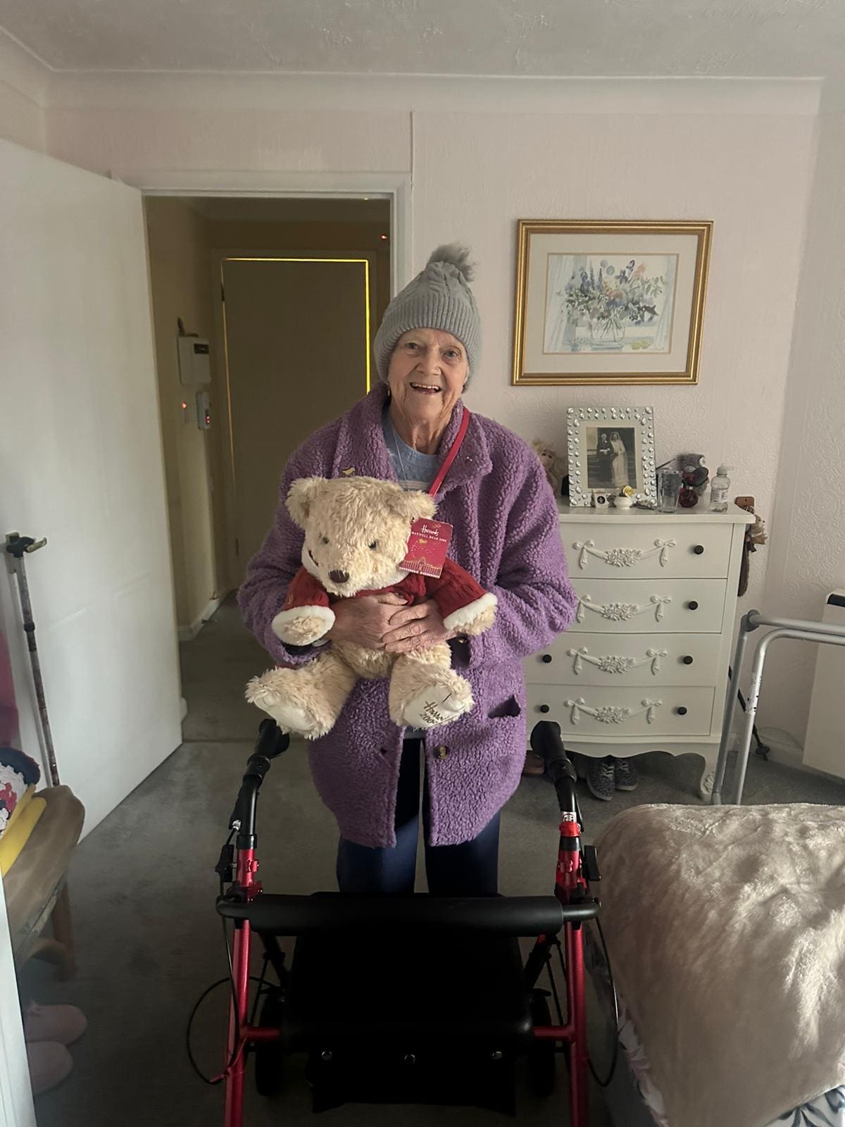 An elderly woman wearing a cozy purple robe and a knitted hat smiles brightly while holding a stuffed teddy bear. She stands beside her walker, showcasing a warm and cheerful atmosphere in her home. This image effectively represents care for elderly individuals, particularly in dementia care, highlighting the importance of comfort, companionship, and warmth during winter months. Providing a nurturing environment and engaging activities can significantly enhance their emotional well-being and overall quality of life for those with dementia.