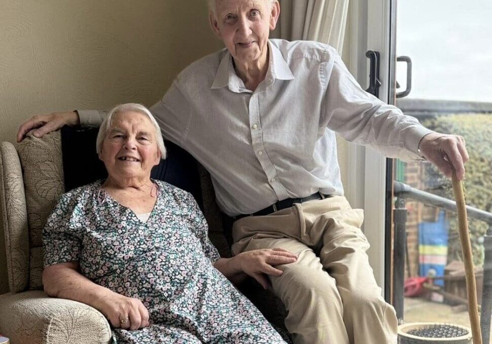 An elderly couple sits together on a cozy sofa, smiling happily. The woman, wearing a floral dress, rests her hand on the man's knee. He holds a cane in his right hand while sitting comfortably beside her. Sunlight streams through a nearby window, highlighting their warm expressions and the inviting atmosphere of their home. This image reflects the importance of companionship and regular check-ups in preventative healthcare, emphasizing how these relationships contribute to overall well-being.