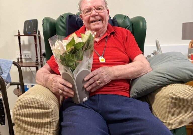 An elderly man sitting in a cozy armchair smiles broadly while holding a bouquet of flowers, exemplifying the joy of receiving thoughtful gestures during the holiday season. Surrounded by a warm and inviting living space, this image highlights the importance of Christmas care and elderly care, showcasing how companionship and small acts of kindness can enhance the well-being of seniors. The cheerful atmosphere reflects the spirit of care and connection that makes the holidays special for older adults.