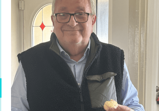 Smiling older man in a vest holding a cupcake and a cupcake liner, standing in a bright kitchen. This image reflects community support through shared moments and connections.