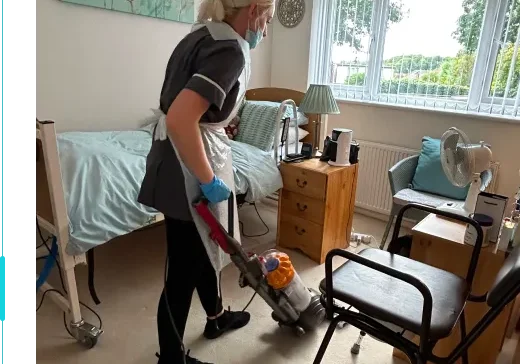 A caregiver begins care by vacuuming a tidy bedroom, wearing gloves and a mask for safety. The organized room features a neat bed and natural light.